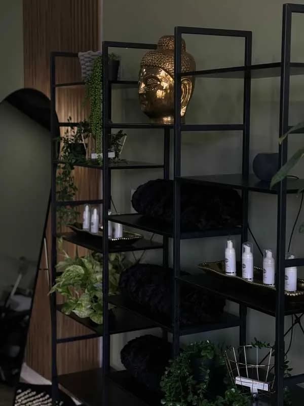 Dimly lit retail shelving at the Lash Addict Studio Clearwater location, styled with a gold Buddha-head accent, rolled towels, and skincare products.