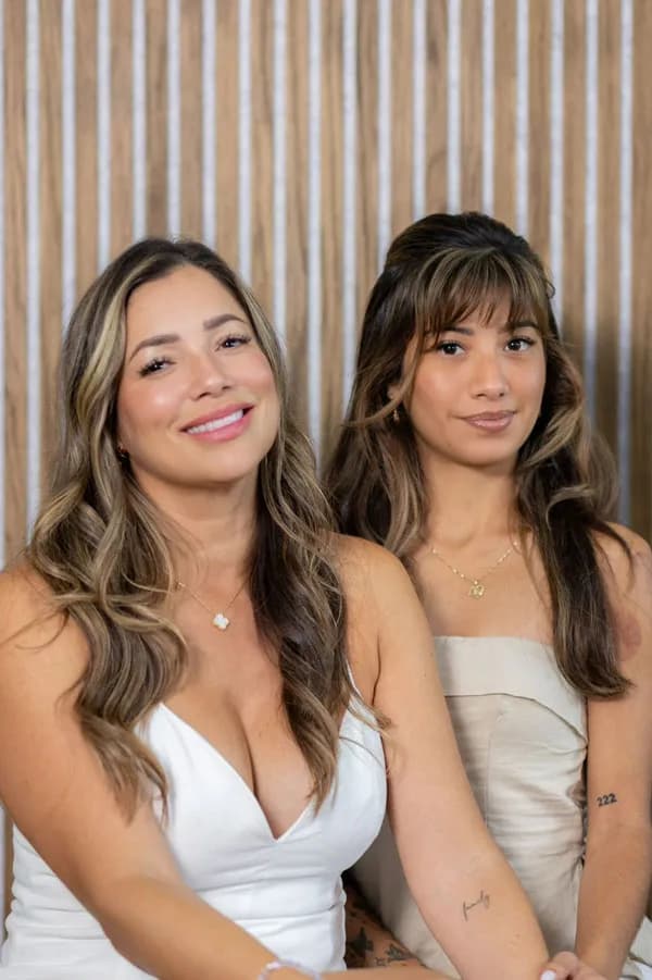Portrait of Natali and Estela, owners of the Lash Addict Studio Seminole location, seated together against a wood-slat wall.