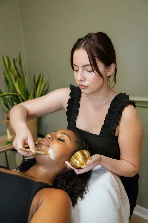 A Lash Addict Studio esthetician applies a facial treatment to a client with a brush and gold mixing bowl.