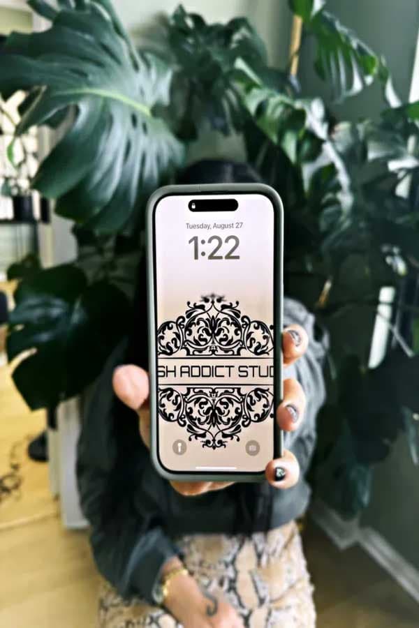 A hand holds up a smartphone showing a Lash Addict Studio lock-screen wallpaper in front of potted plants.